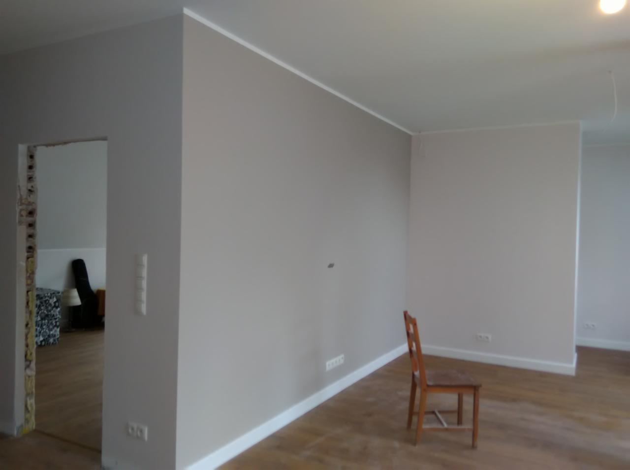 Wnętrze po remoncie: ściany pomalowane na szaro, białe listwy przypodłogowe i sufitowe, widoczne gniazdka elektryczne, drewniana podłoga, krzesło i częściowo wykończone przejście między...