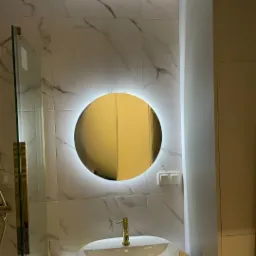 Nowoczesna łazienka: okrągłe lustro z podświetleniem LED nad owalną umywalką z złotą baterią na tle płytek imitujących marmur. Minimalistyczny design i eleganckie wykończenie.