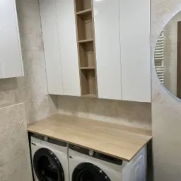 Nowoczesna łazienka z zabudową meblową na wymiar: białe szafki wiszące z otwartą półką i drewniany blat nad pralkami. Minimalistyczny design, beżowe płytki.