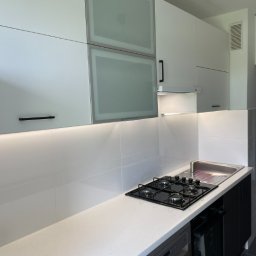 DJK Meble Sp. z o.o. - Nowoczesna, biała kuchnia z czarnymi akcentami w Krakowie: minimalistyczne szafki, podświetlany blat, płyta gazowa i zlew w jednym ciągu. Ujęcie z bliska.