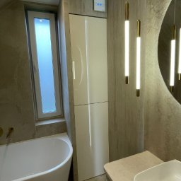 DJK Meble Sp. z o.o. - Nowoczesna łazienka z białą szafką, wanną i wiszącymi lampami. Uwagę przyciąga okrągłe lustro i płytki imitujące kamień w neutralnych odcieniach.