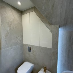 DJK Meble Sp. z o.o. - Nowoczesna łazienka w beżowych tonacjach z podwieszaną toaletą, bidetem z złotą armaturą i szafką wnękową pod skosem. Stonowana kolorystyka i minimalistyczny design.