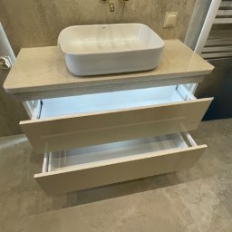 DJK Meble Sp. z o.o. - Nowoczesna łazienka: biała umywalka nablatowa, złota armatura i szafka z szufladami z podświetleniem LED, na tle beżowych płytek imitujących kamień.