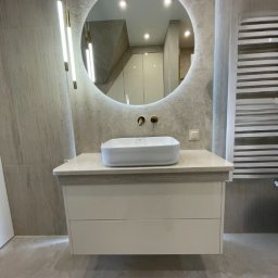 DJK Meble Sp. z o.o. - Nowoczesna łazienka z okrągłym lustrem LED, białą szafką pod umywalkę i dekoracyjnym grzejnikiem. Minimalistyczny design z szarymi płytkami i wiszącymi lampami.