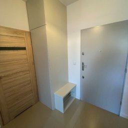 DJK Meble Sp. z o.o. - Wnętrze przedpokoju: wysoka szafa w zabudowie w odcieniach szarości, drewniane drzwi po lewej, szare drzwi wejściowe po prawej, biała szafka z półkami.