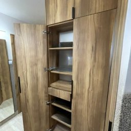 DJK Meble Sp. z o.o. - Otwarta szafa z drewna o wyraźnym usłojeniu, z półkami i szufladą. Widoczne metalowe zawiasy i podłużne, czarne uchwyty. W tle fragment lustra i jasna podłoga.