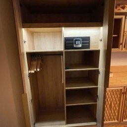 DJK Meble Sp. z o.o. - Drewniana szafa z otwartymi drzwiami, widoczne półki, wieszaki i sejf. Ujęcie z bliska, oświetlenie wnętrza szafy podkreśla strukturę drewna.