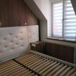 DJK Meble Sp. z o.o. - Białe łóżko z tapicerowanym zagłówkiem w pokoju na poddaszu, z oknem z żaluzjami, oświetleniem ściennym i brązową boazerią. Ujęcie z dołu.