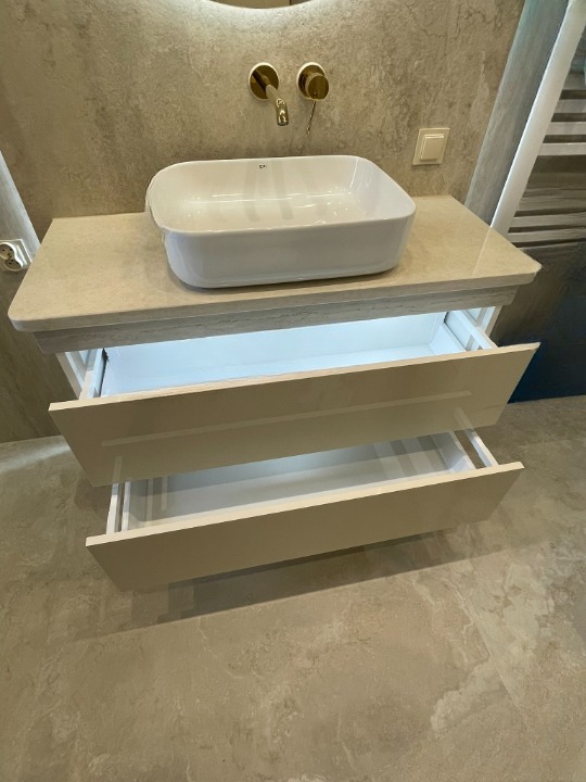 Nowoczesna łazienka: biała umywalka nablatowa, złota armatura i szafka z szufladami z podświetleniem LED, na tle beżowych płytek imitujących kamień.