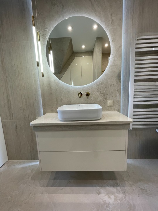 Nowoczesna łazienka z okrągłym lustrem LED, białą szafką pod umywalkę i dekoracyjnym grzejnikiem. Minimalistyczny design z szarymi płytkami i wiszącymi lampami.