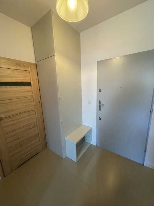 Wnętrze przedpokoju: wysoka szafa w zabudowie w odcieniach szarości, drewniane drzwi po lewej, szare drzwi wejściowe po prawej, biała szafka z półkami.