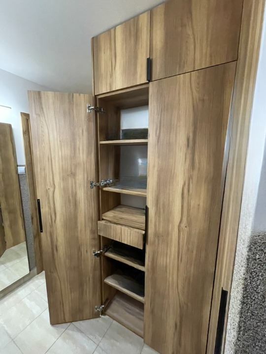 Otwarta szafa z drewna o wyraźnym usłojeniu, z półkami i szufladą. Widoczne metalowe zawiasy i podłużne, czarne uchwyty. W tle fragment lustra i jasna podłoga.