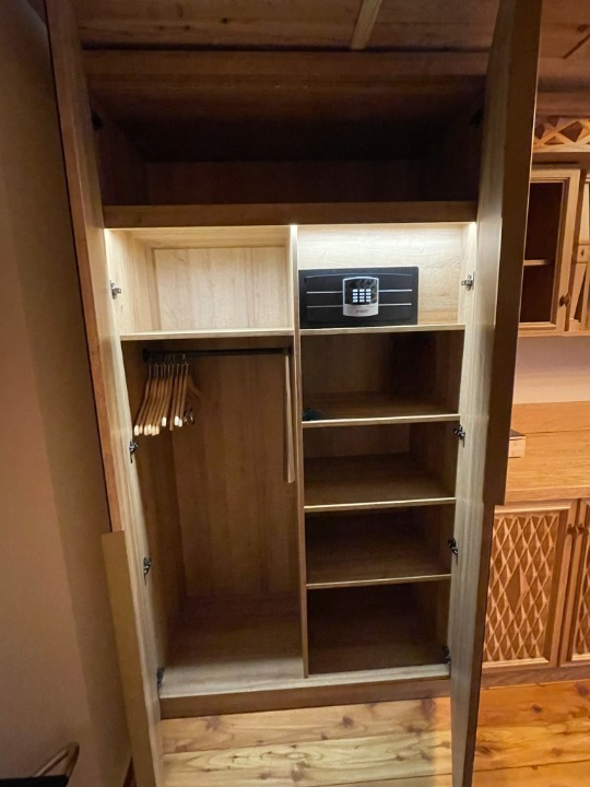 Drewniana szafa z otwartymi drzwiami, widoczne półki, wieszaki i sejf. Ujęcie z bliska, oświetlenie wnętrza szafy podkreśla strukturę drewna.