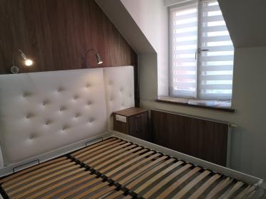 Białe łóżko z tapicerowanym zagłówkiem w pokoju na poddaszu, z oknem z żaluzjami, oświetleniem ściennym i brązową boazerią. Ujęcie z dołu.