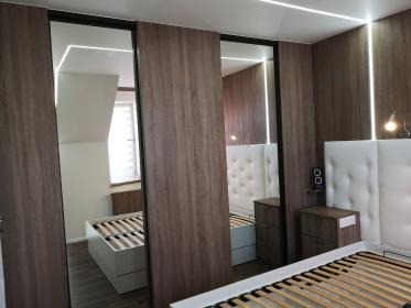Szafa wnękowa z lustrzanymi drzwiami przesuwnymi, imitacja drewna, oświetlenie LED, wnętrze sypialni z łóżkiem i szafką nocną. Minimalistyczny design.