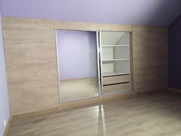 Jasna szafa wnękowa z lustrzanymi drzwiami przesuwnymi, dopasowana do skosu poddasza, z widocznymi półkami i szufladami. Parkiet w odcieniach beżu.