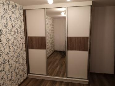 Szafa wnękowa z przesuwnymi drzwiami: białe panele z poziomym pasem w kolorze drewna i lustrem pośrodku. Wnętrze z wzorzystą tapetą i drewnianą podłogą.