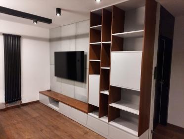 Nowoczesna szafa wnękowa z drewnianymi półkami i białymi frontami, wkomponowana w szarą ścianę z telewizorem, na tle drewnianej podłogi.
