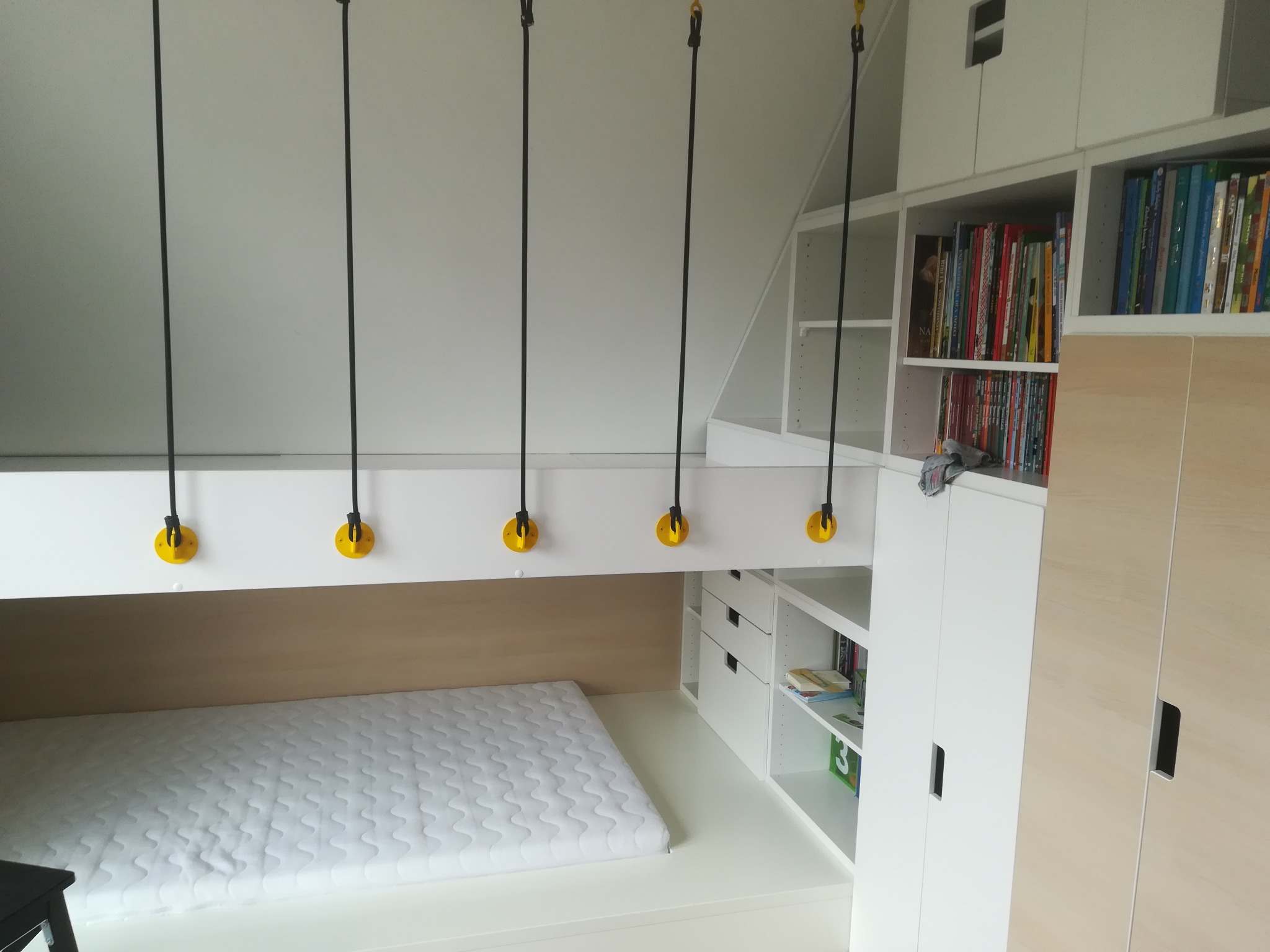 Pokój dziecięcy z łóżkiem na antresoli, białe meble z półkami na książki i szufladami, czarne liny z żółtymi elementami mocującymi do sufitu, biały materac na łóżku.