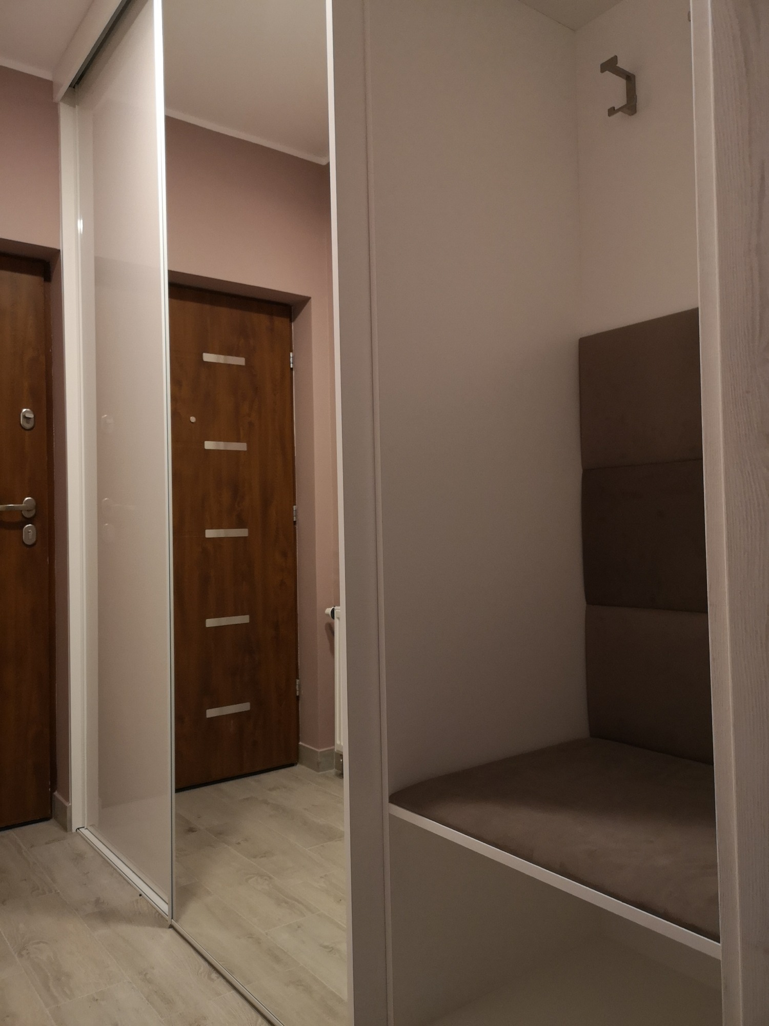 Wnętrze przedpokoju z białą szafą przesuwną z lustrem, w której odbija się brązowe drzwi wejściowe, oraz siedziskiem z tapicerowanym oparciem i wieszakiem na ścianie.