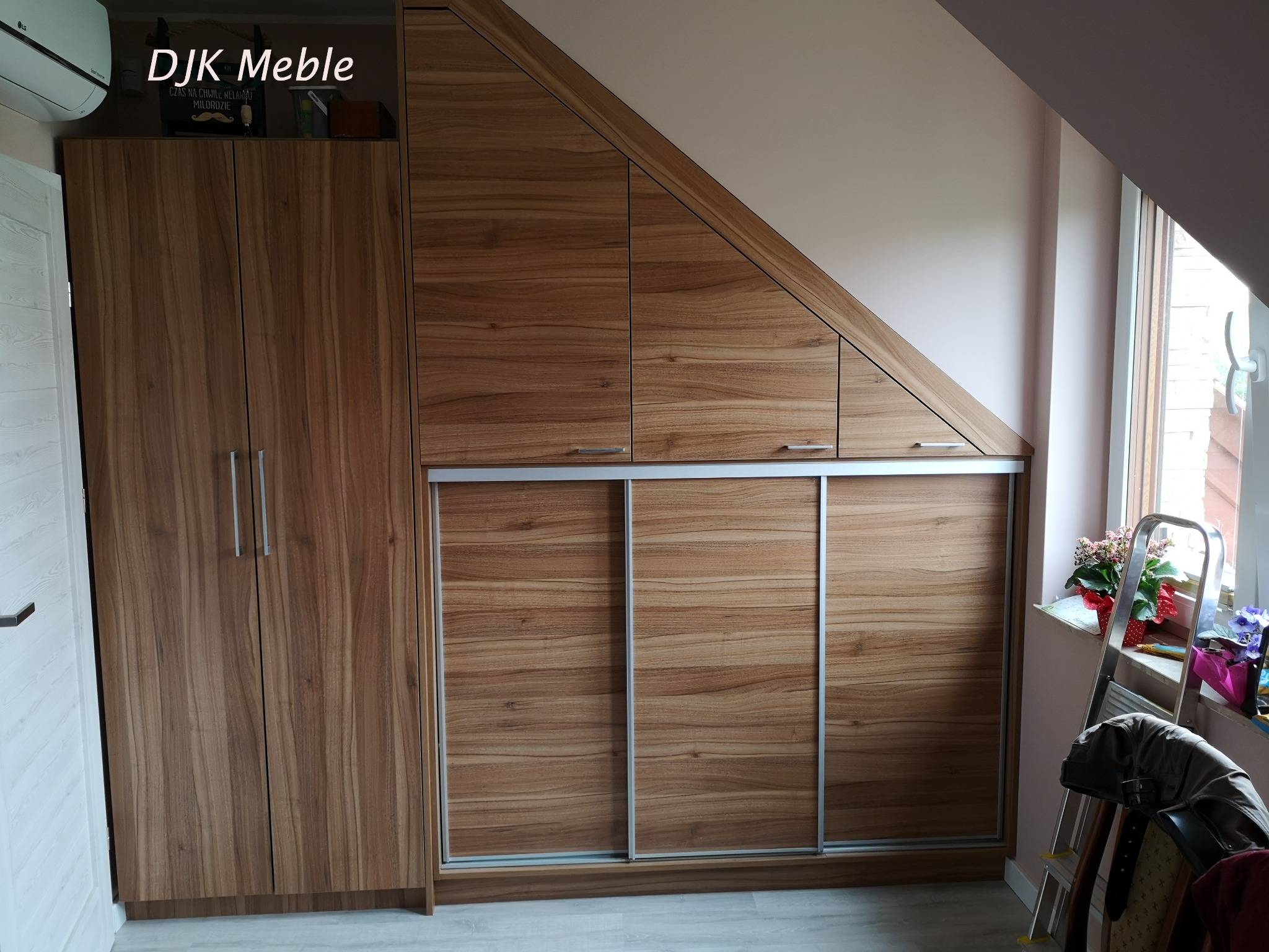 Drewniana szafa z przesuwnymi drzwiami i szafkami pod skosem na poddaszu, widoczny fragment drabiny i kwiaty na parapecie.