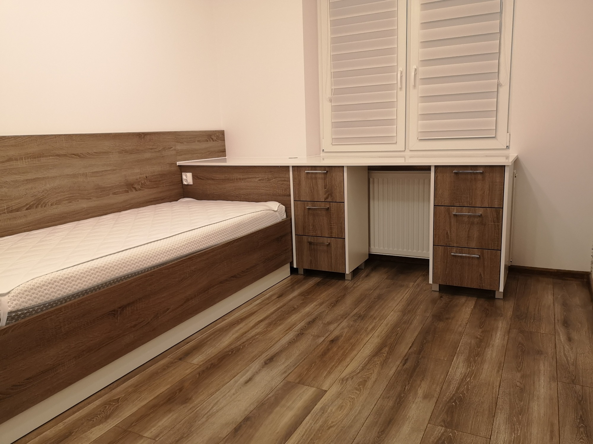 Aranżacja pokoju z łóżkiem i biurkiem z szafkami w zabudowie okiennej, wykończone w dekorze drewna z białymi elementami, na podłodze panele imitujące drewno.