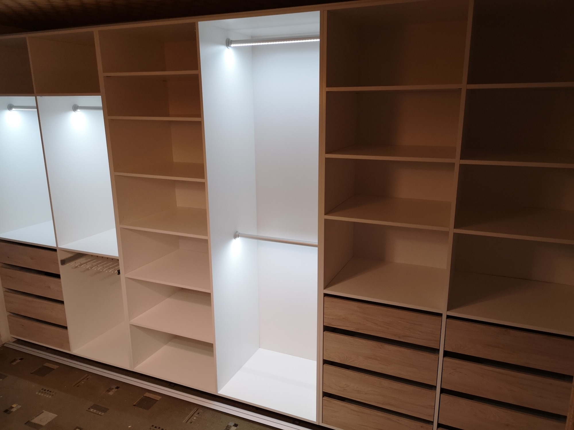 Przestronna garderoba z białymi półkami i drewnianymi szufladami, oświetlona listwami LED, z widocznym systemem przesuwnym na podłodze.