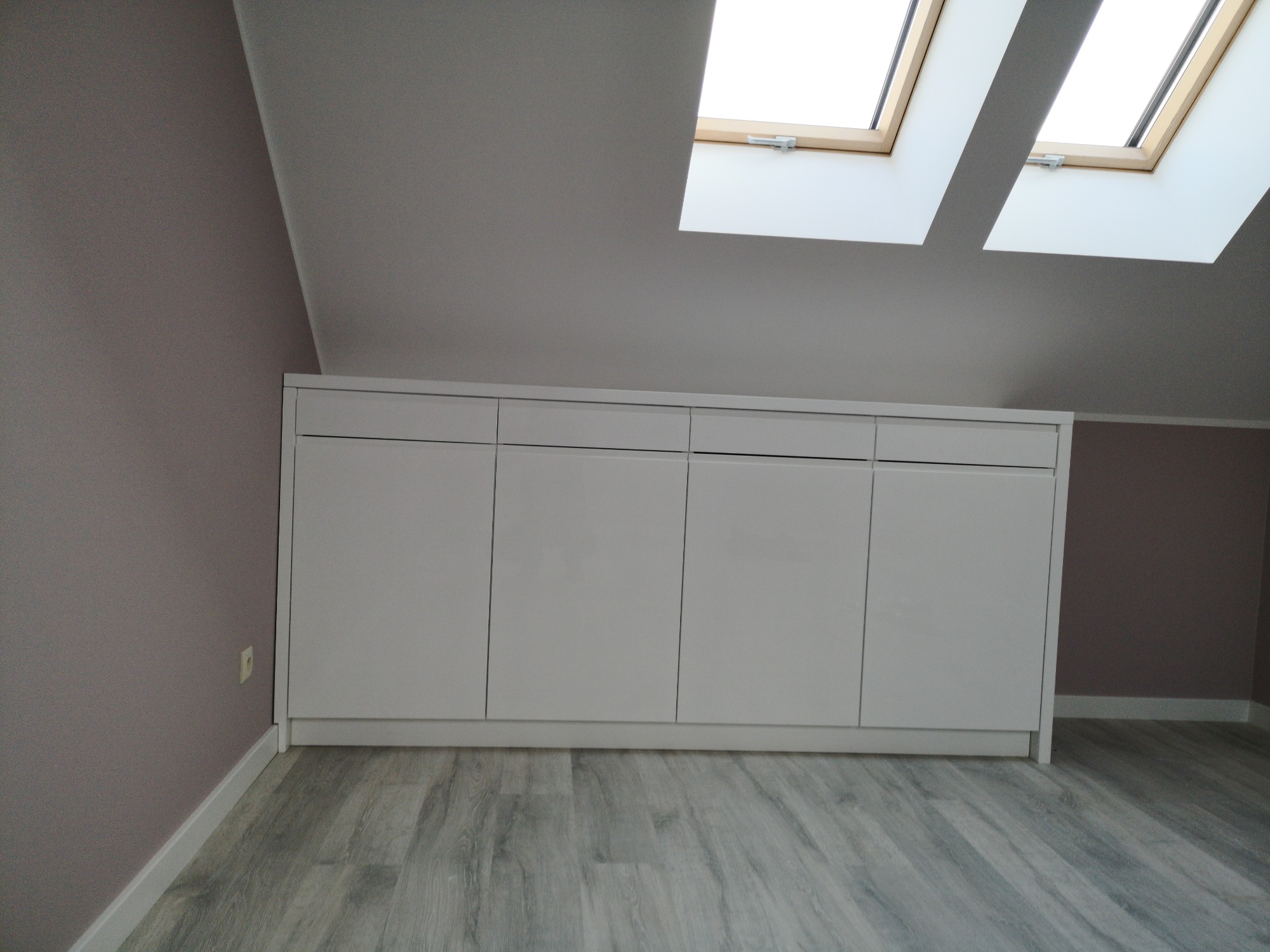 Biała szafa z czterema drzwiami, zamontowana pod skosem dachu, z dwoma oknami dachowymi, na podłodze panele imitujące drewno.