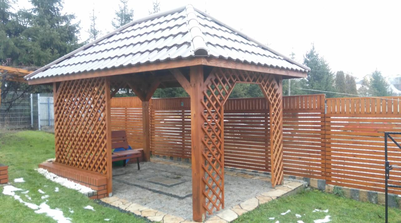 Drewniana altana ogrodowa z kratowanymi ścianami, kryta blachodachówką, z ławką wewnątrz i fragmentami śniegu na trawniku.