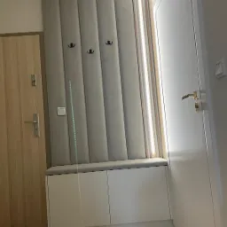 Szara tapicerowana ściana z wieszakami i siedziskiem nad białą szafką w przedpokoju, obok drzwi wejściowych i wewnętrznych.