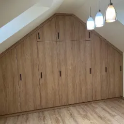 Szafa wnękowa z drewnianymi frontami, dopasowana do skosu dachu w Pcimiu, z czarnymi, minimalistycznymi uchwytami, oświetlona trzema wiszącymi lampami.