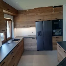 Nowoczesna kuchnia z drewnianymi frontami szafek, czarnym blatem i dużym, czarnym lodówko-zamrażarką side-by-side; widoczne niedokończone podłączenia oświetlenia sufitowego.