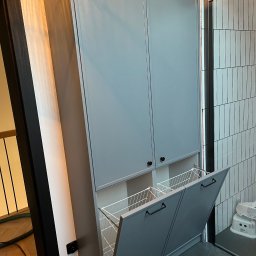 FIRMA USŁUGOWO-HANDLOWA ALINA ULMAN - Szara szafa wnękowa z koszami na pranie, otwarte drzwiczki, czarne uchwyty, minimalistyczny design, płytki na ścianie w tle, nowoczesne wykończenie.