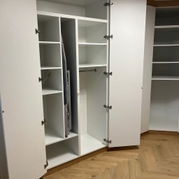 FIRMA USŁUGOWO-HANDLOWA ALINA ULMAN - Biała szafa wnękowa z otwartymi drzwiami, widoczne półki, drążek na ubrania i deska do prasowania. Parkiet w jodełkę na podłodze. Oświetlenie sufitowe.