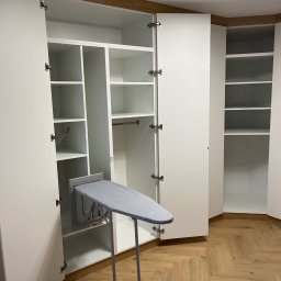 FIRMA USŁUGOWO-HANDLOWA ALINA ULMAN - Biała szafa wnękowa z otwartymi drzwiami, wewnątrz półki i wysuwana deska do prasowania. Parkiet w jodełkę na podłodze, nowoczesne oświetlenie sufitowe.