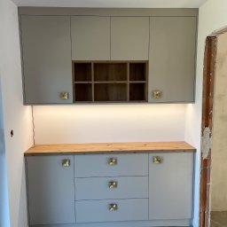 FIRMA USŁUGOWO-HANDLOWA ALINA ULMAN - Jasnoszara zabudowa kuchenna z drewnianym blatem i złotymi uchwytami, oświetlona ledami, na tle surowego muru. Widoczne trzy szuflady i szafki wiszące.