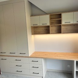 FIRMA USŁUGOWO-HANDLOWA ALINA ULMAN - Jasna zabudowa meblowa z szafą, szafkami wiszącymi i biurkiem z blatem drewnopodobnym, czarne uchwyty, oświetlenie LED pod szafkami wiszącymi.