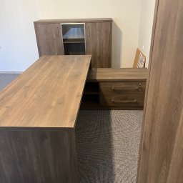 FIRMA USŁUGOWO-HANDLOWA ALINA ULMAN - Kątowe biurko z szafkami w odcieniach brązu, z widoczną strukturą drewna, na tle szarej wykładziny. Elementy biurowe w tle.