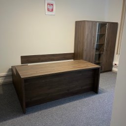 FIRMA USŁUGOWO-HANDLOWA ALINA ULMAN - Biuro z ciemnobrązowym biurkiem i szafą z przeszkleniami, na ścianie godło Polski. Wykładzina dywanowa w szarym odcieniu. Oświetlenie sufitowe.