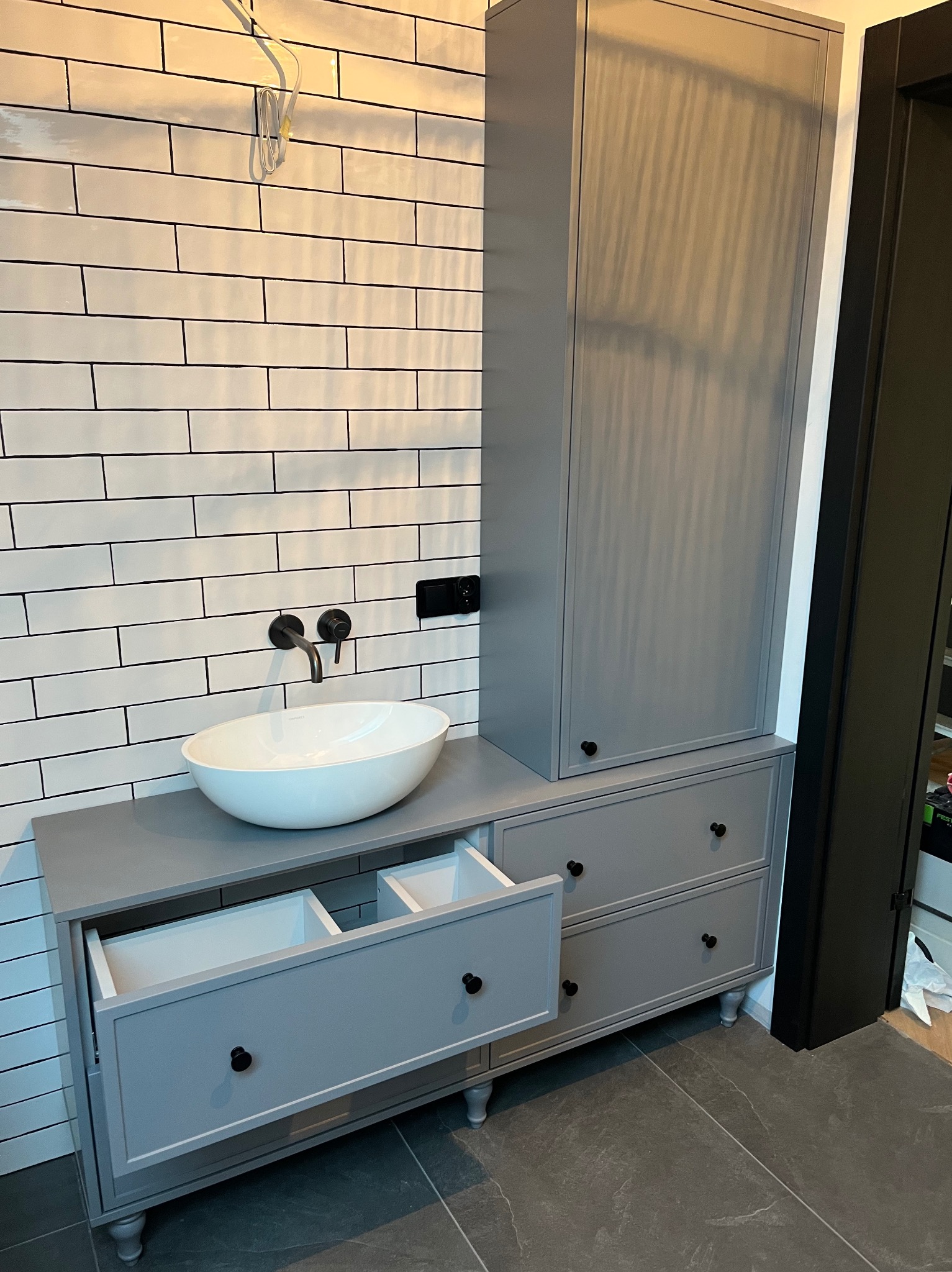 Szara szafka łazienkowa z umywalką nablatową i otwartymi szufladami na tle białych płytek imitujących cegłę. Nowoczesny design z czarną armaturą.
