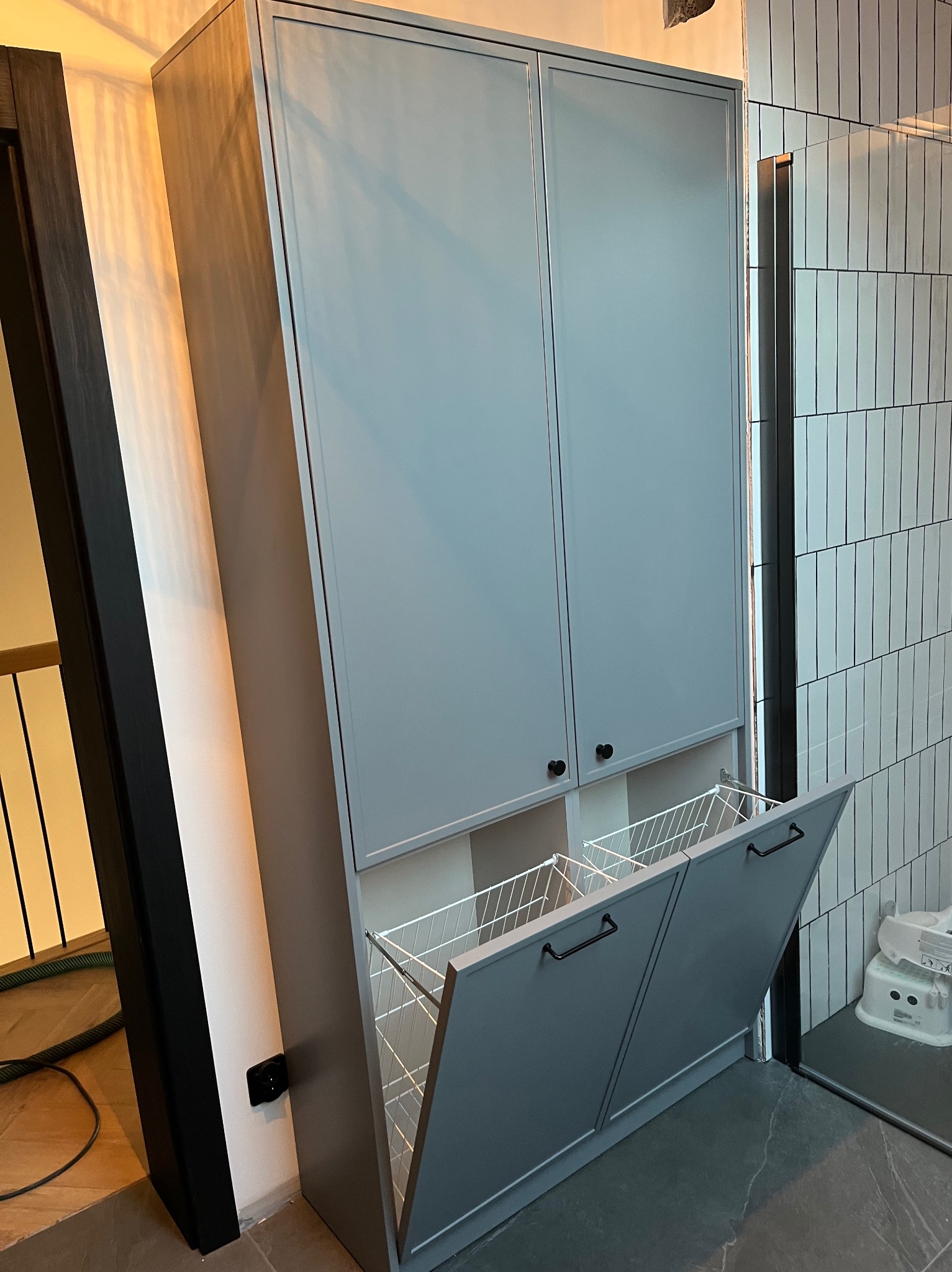 Szara szafa wnękowa z koszami na pranie, otwarte drzwiczki, czarne uchwyty, minimalistyczny design, płytki na ścianie w tle, nowoczesne wykończenie.