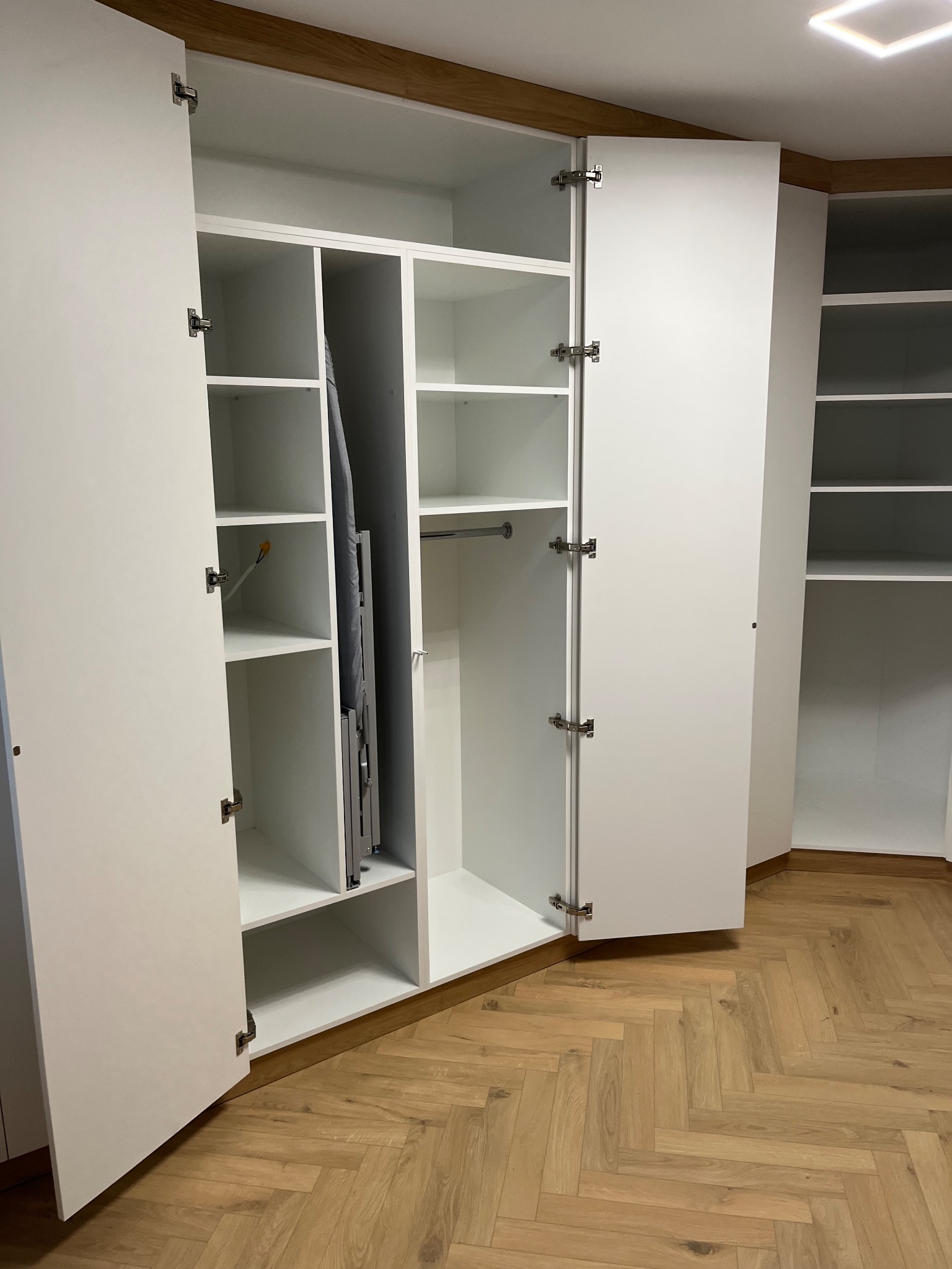Biała szafa wnękowa z otwartymi drzwiami, widoczne półki, drążek na ubrania i deska do prasowania. Parkiet w jodełkę na podłodze. Oświetlenie sufitowe.