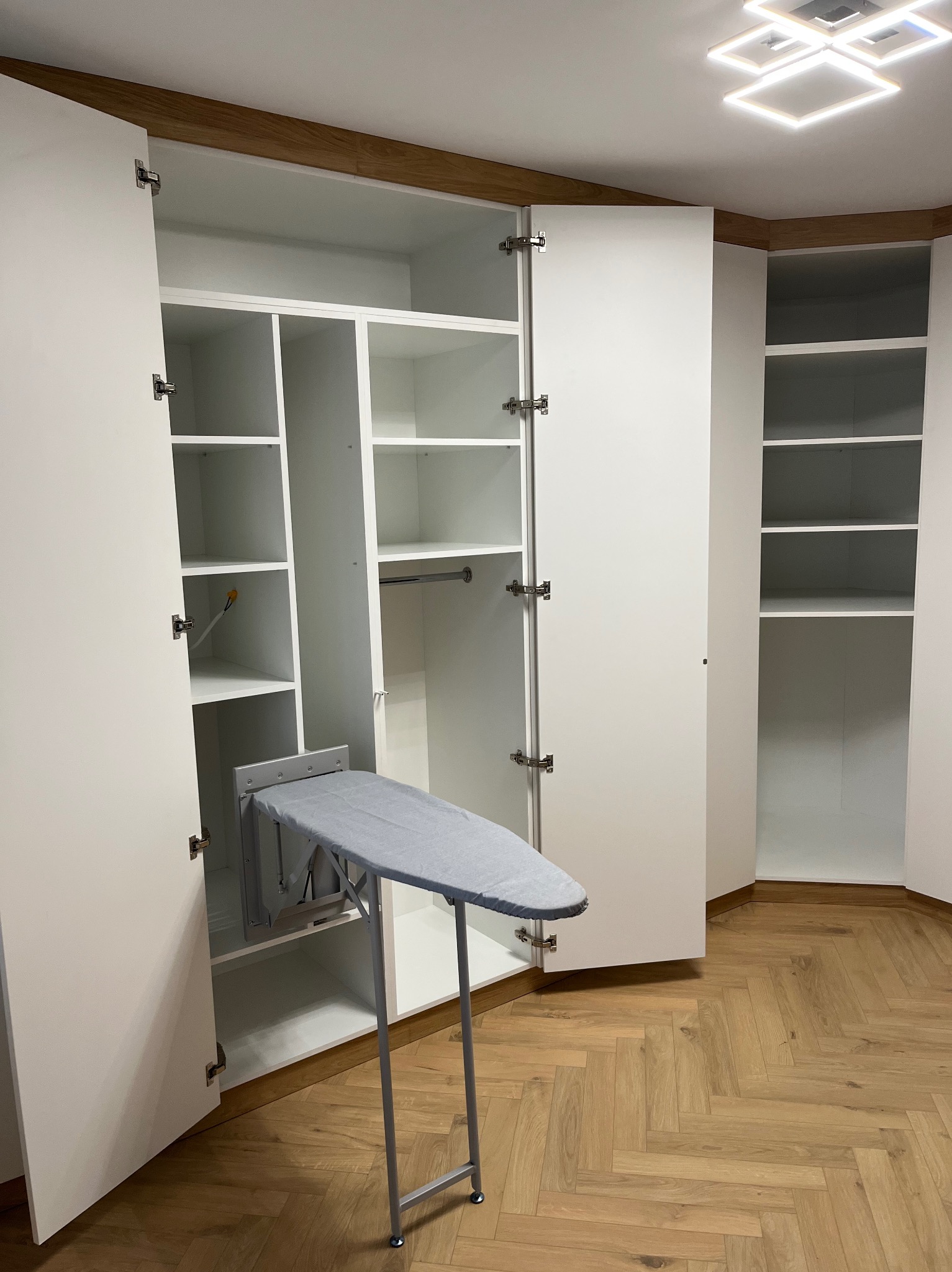 Biała szafa wnękowa z otwartymi drzwiami, wewnątrz półki i wysuwana deska do prasowania. Parkiet w jodełkę na podłodze, nowoczesne oświetlenie sufitowe.