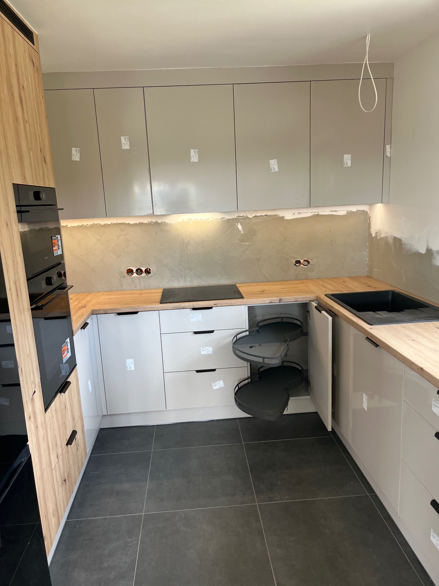 Jasna kuchnia z drewnianym blatem i szafką narożną z półkami obrotowymi. Widoczne niedokończone wykończenie ściany nad blatem. Nowoczesny design.