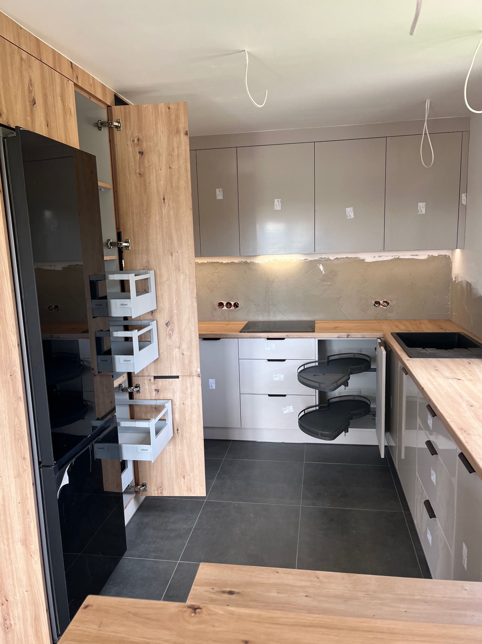 Nowoczesna kuchnia z drewnianymi frontami i szarymi szafkami. Otwarta szafka narożna z półkami obrotowymi i wysuwane cargo Blum. Widoczne gniazdka elektryczne.