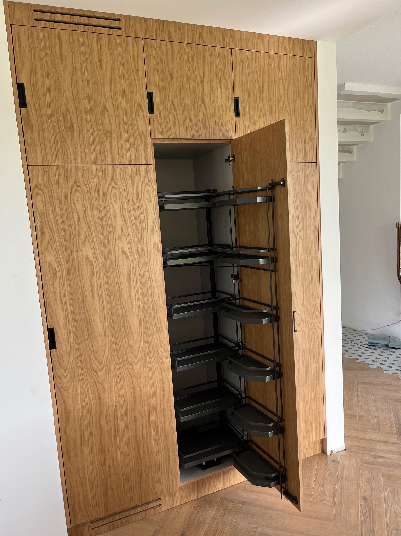 Drewniana szafa na wymiar z systemem cargo, otwarte drzwi ukazują półki. Parkiet w jodełkę na podłodze. Minimalistyczny design, czarne uchwyty.