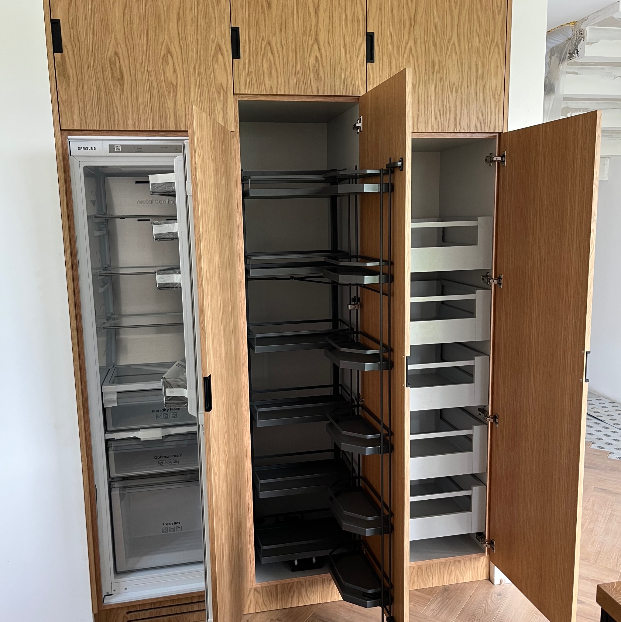 Drewniana zabudowa kuchenna z otwartymi szafkami, widoczny system cargo i szuflady. Wkomponowana lodówka Samsung. Jasne drewno, minimalistyczne uchwyty.