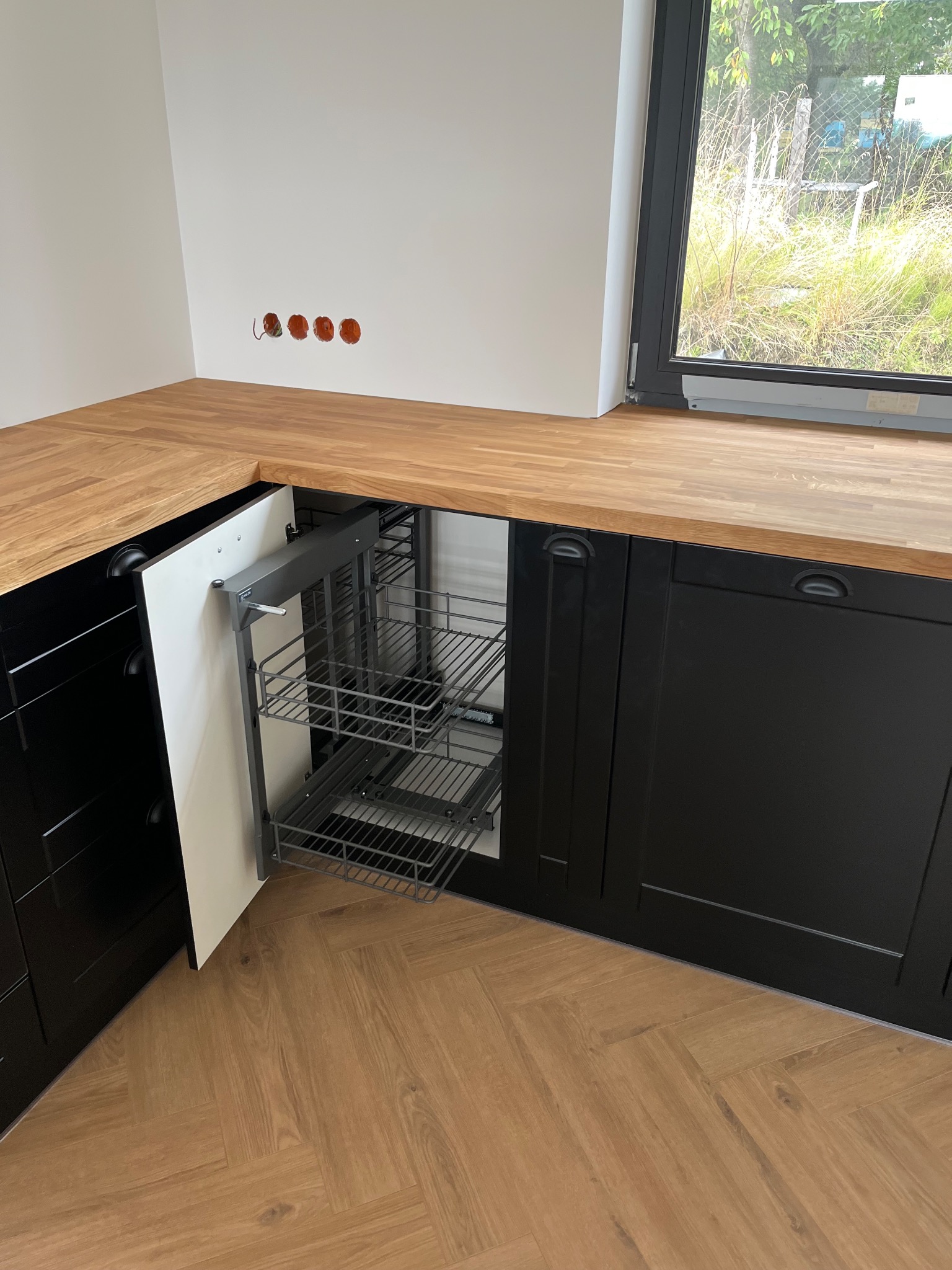 Czarne szafki kuchenne z drewnianym blatem i wysuwanym koszem cargo w rogu. Widoczna podłoga z paneli ułożonych w jodełkę i okno z widokiem na zieleń.