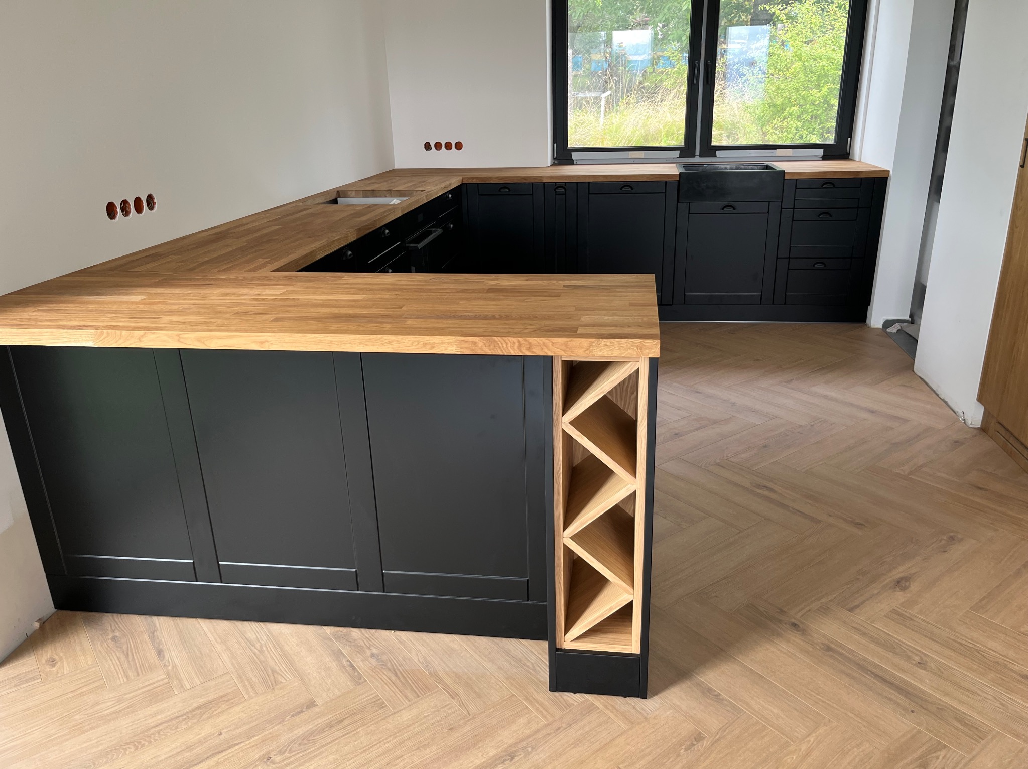 Nowoczesna kuchnia w kształcie litery U z czarnymi frontami i drewnianym blatem. Widoczny regał na wino i fragment podłogi ułożonej w jodełkę. Surowy stan ścian.