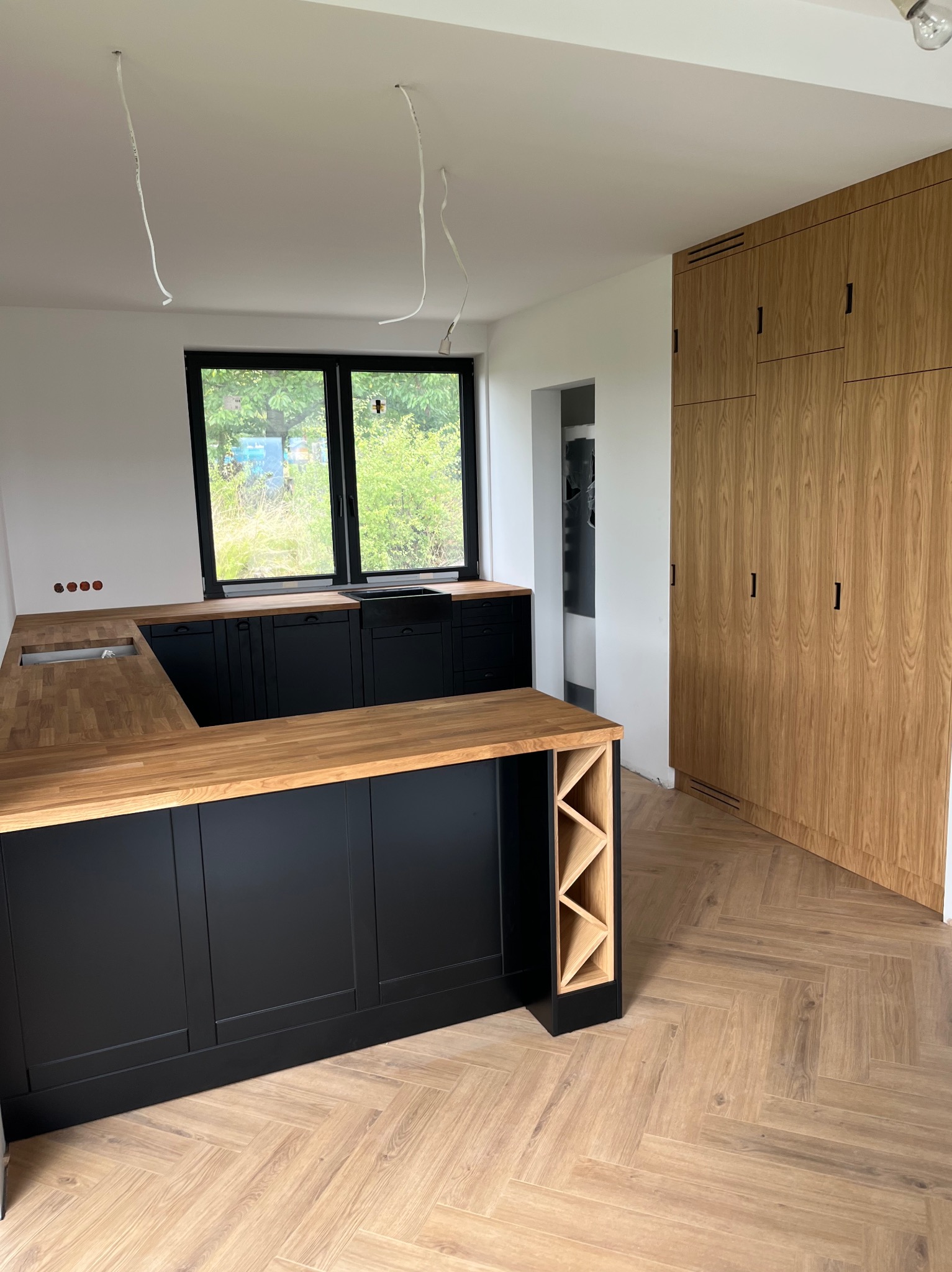 Nowoczesna kuchnia: czarne szafki z drewnianym blatem, szafa w zabudowie z forniru dębowego, parkiet w jodełkę. Widoczne kable oświetleniowe.
